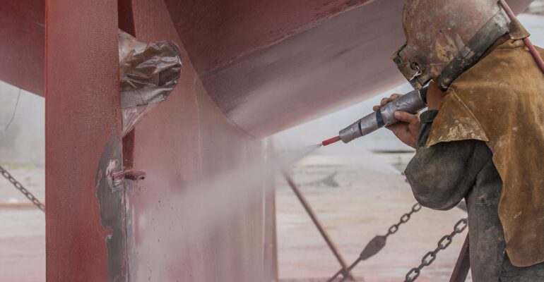 worker, sandblasting the corroded hull of a sailing vessel with a high pressure sandblasting syst