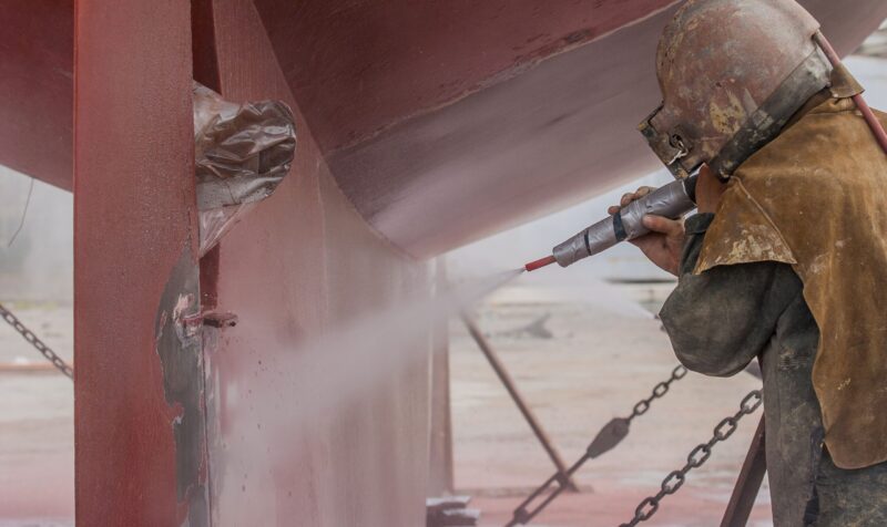 worker, sandblasting the corroded hull of a sailing vessel with a high pressure sandblasting syst