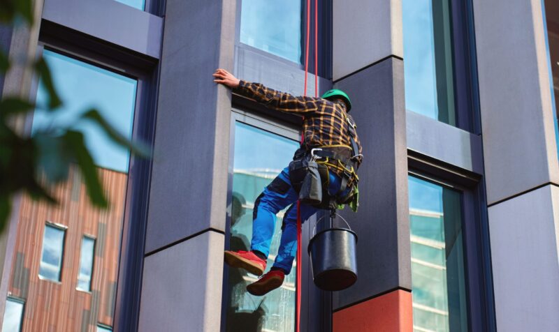 Window cleaner cleaning glass facade of tall modern office building with safety harness, bucket and climbing equipment. Concept of maintenance, safety and urban exterior services